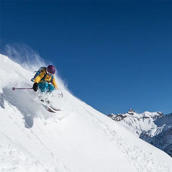 Een skiër in een geel jack en een paarse helm snijdt door de verse poedersneeuw op een steile, besneeuwde helling, omringd door met sneeuw bedekte bergen onder een strakblauwe hemel.